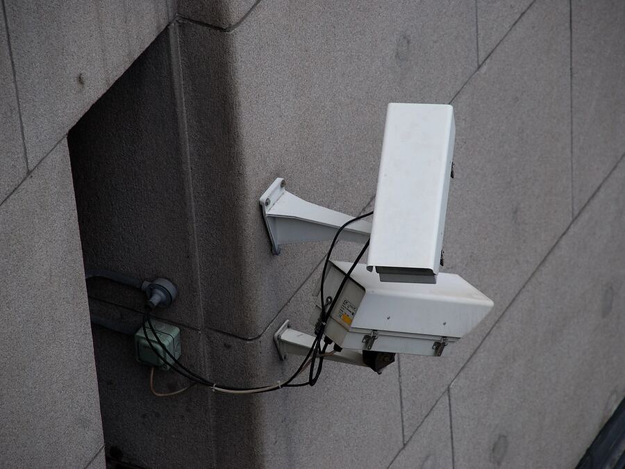 A static CCTV camera bolted to a wall bracket, its cable run fixed along the surface